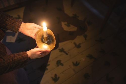 kädet pitelevät kynttilää ja varjoisalle lattialle on levitetty havuja.