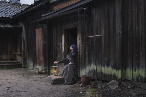 huivipäinen nainen istuu vanhan talon kynnyksellä lyhty kädessä.