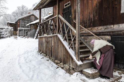 A woman in 19th century clothing sweeps snow on the stairs.